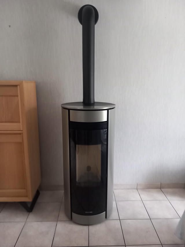 Un grand poêle à bois cylindrique, argenté et noir, posé sur un sol carrelé contre un mur nu.