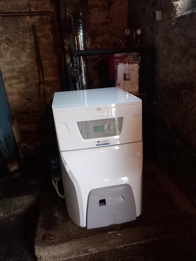 Une chaudière blanche moderne avec un écran numérique, située dans un sous-sol faiblement éclairé, à côté d'un conduit de cheminée.