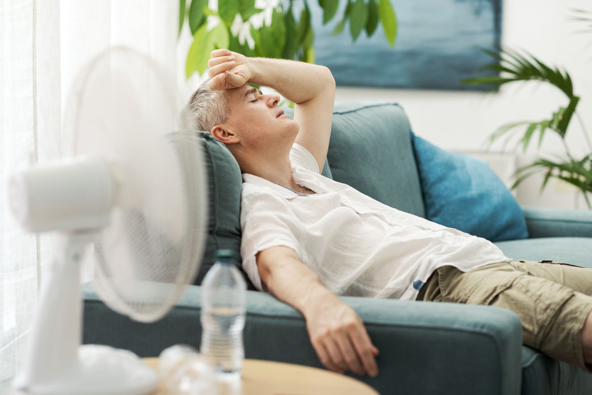 Une personne allongée sur un canapé turquoise dans une pièce lumineuse s'essuie la sueur tandis qu'un ventilateur souffle à proximité.