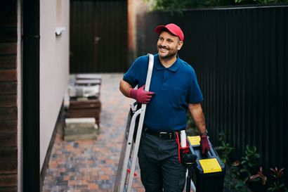 Un ouvrier portant une casquette rouge, des outils et une échelle, sourit à l'extérieur, près d'un bâtiment.