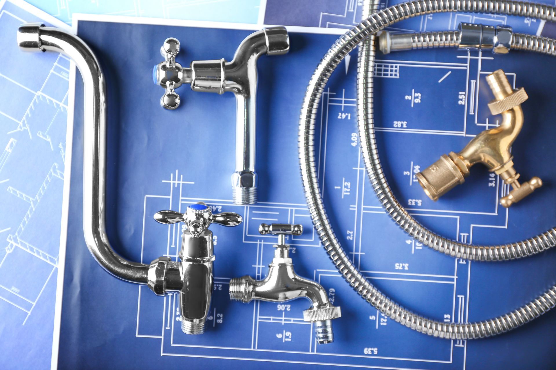 Divers robinets chromés et en laiton, ainsi qu'un flexible de douche en métal, sont disposés sur des plans architecturaux bleus.