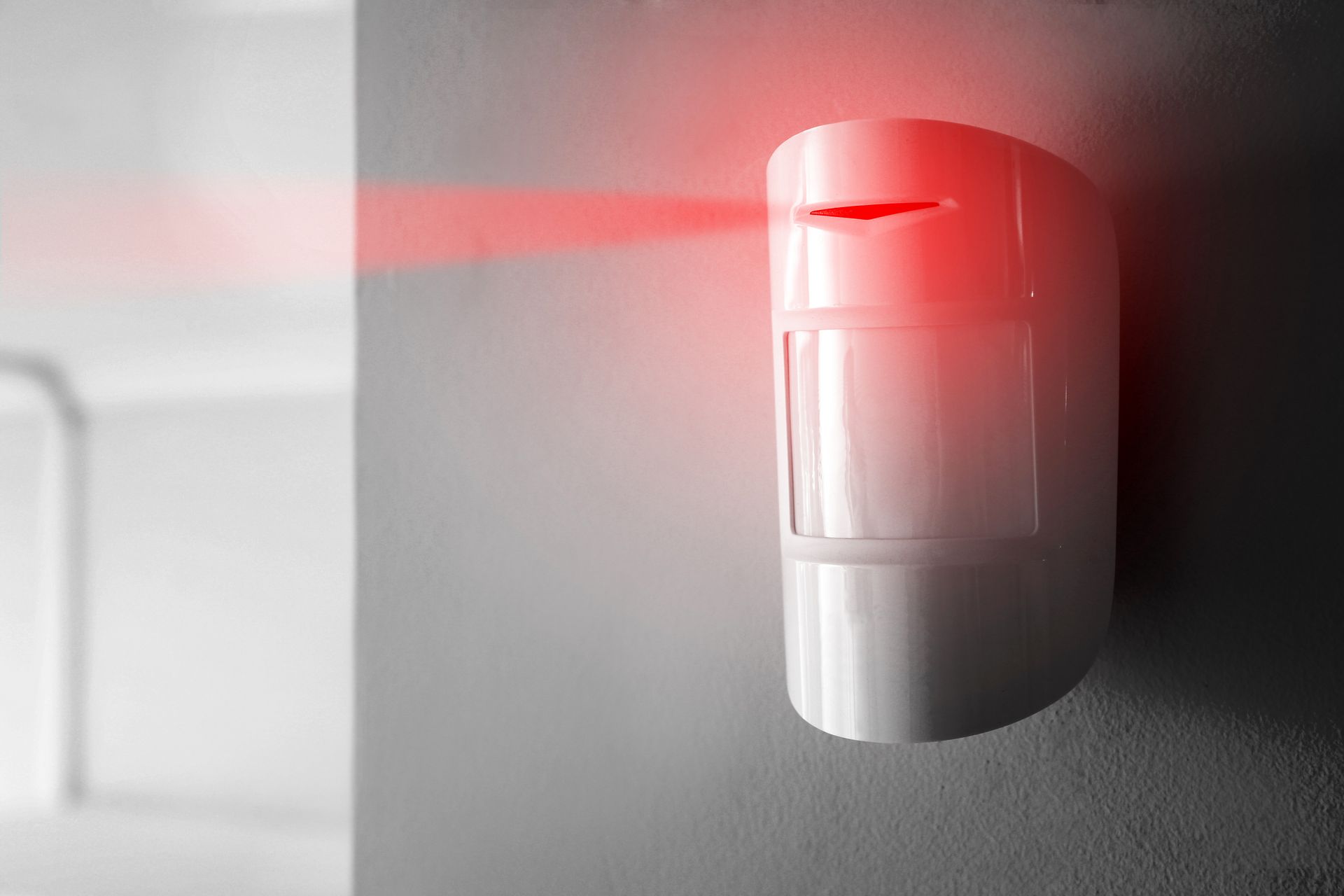 Un détecteur de mouvement blanc fixé au mur, émettant un faisceau laser horizontal rouge vif.