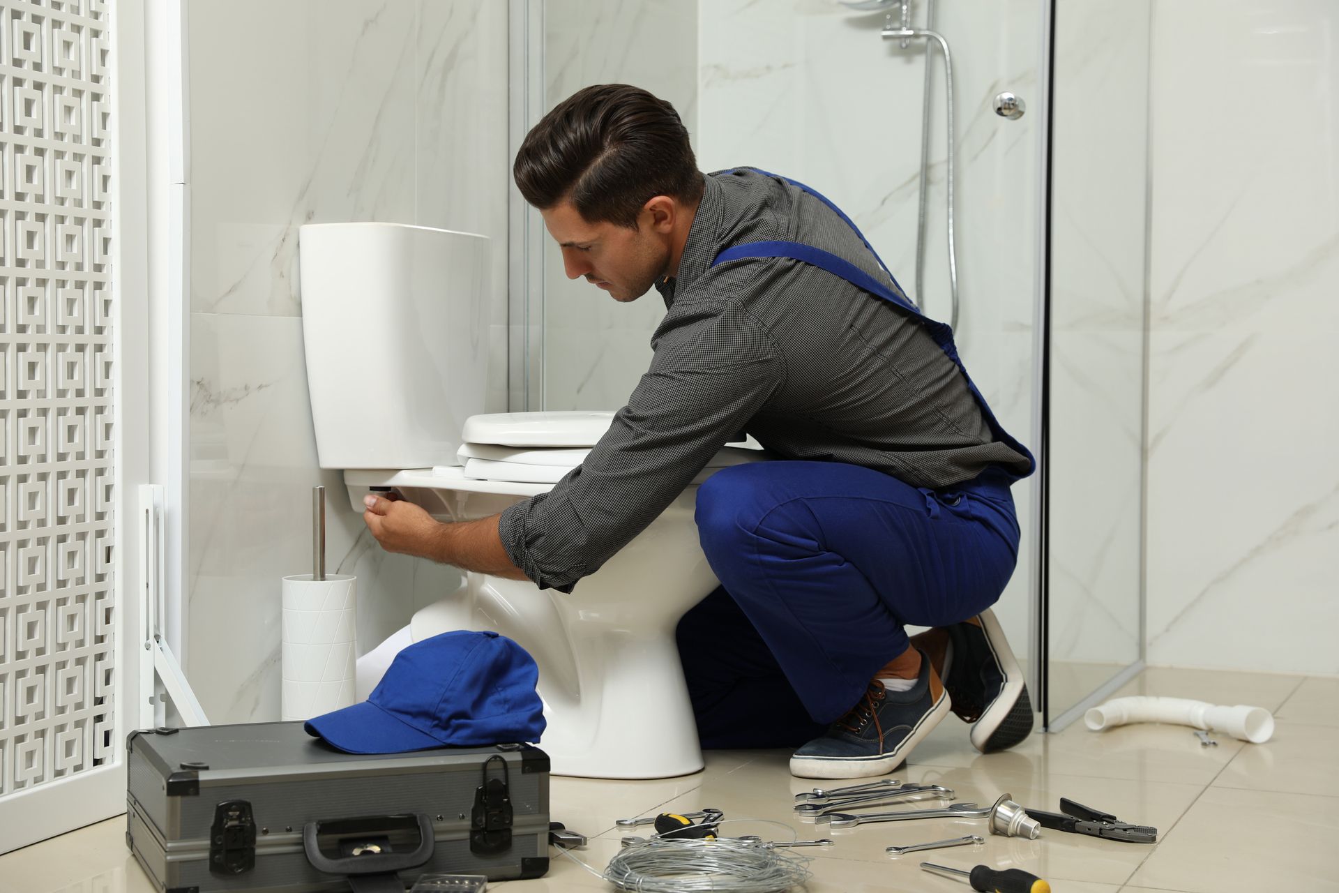Un homme agenouillé près des toilettes, manipulant des outils dans une salle de bains dont des pièces sont éparpillées sur le sol.