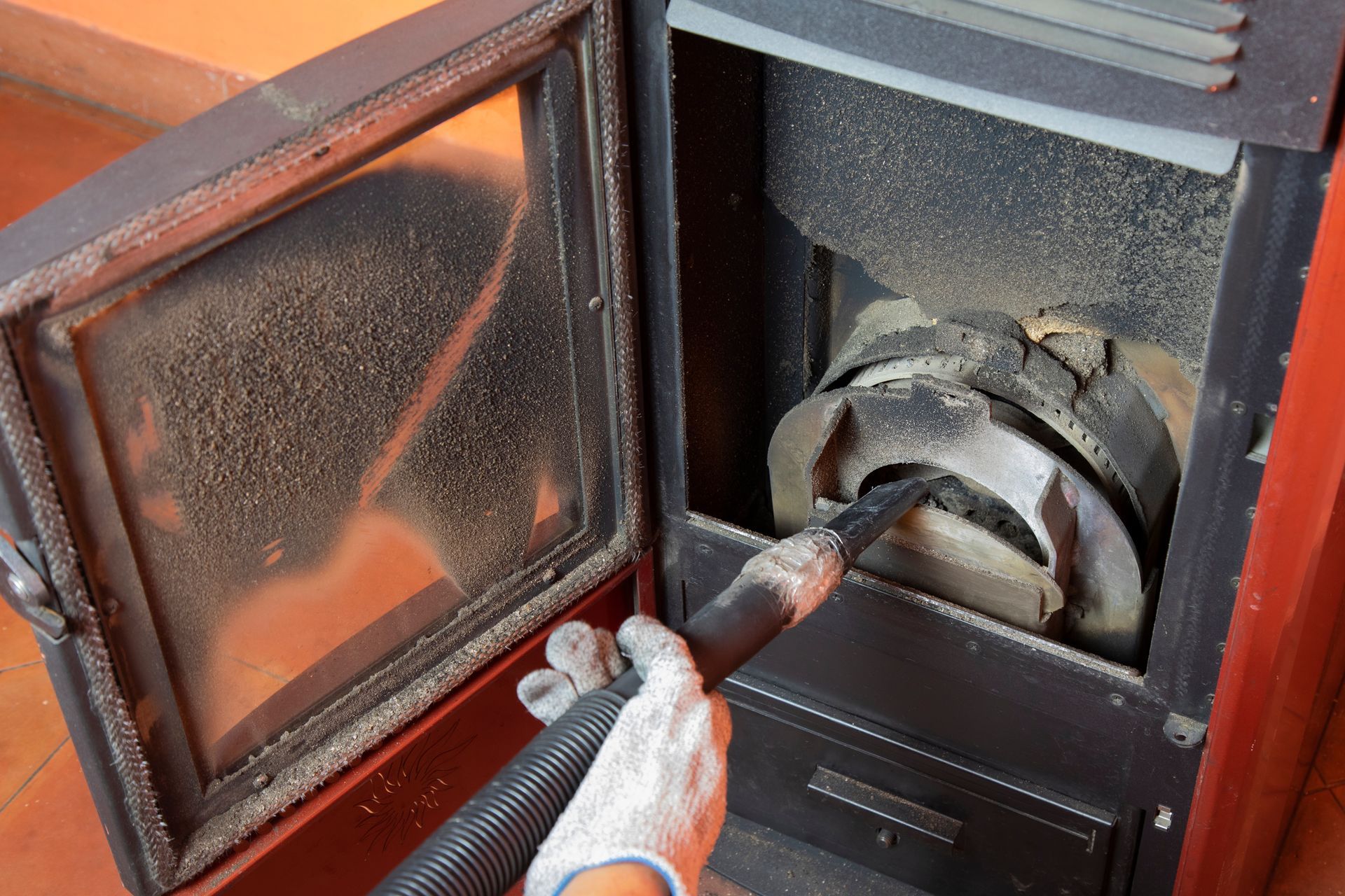 Une personne portant un gant de travail gris utilise un tuyau d'aspirateur pour nettoyer les cendres à l'intérieur d'un poêle à granulés.