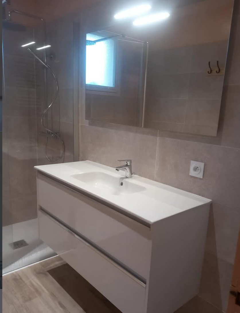 Salle de bains moderne avec meuble-lavabo mural blanc, lavabo carré, grand miroir et douche vitrée sur carrelage beige.