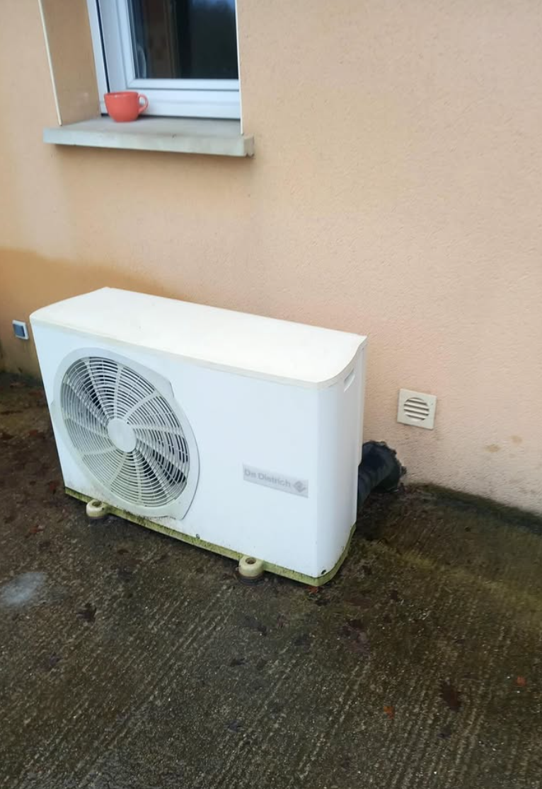 Une pompe à chaleur blanche est installée sur une terrasse en béton à l'extérieur, adossée à un mur clair et placée sous une petite fenêtre.