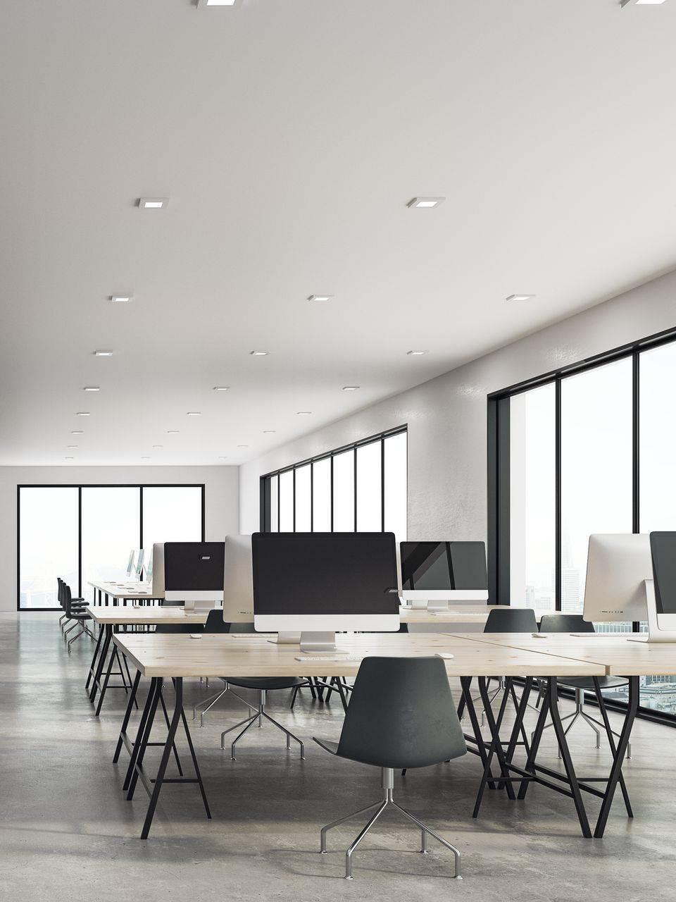 Bureau moderne vide avec de longs bureaux, des ordinateurs et de grandes fenêtres laissant entrer une lumière naturelle abondante.
