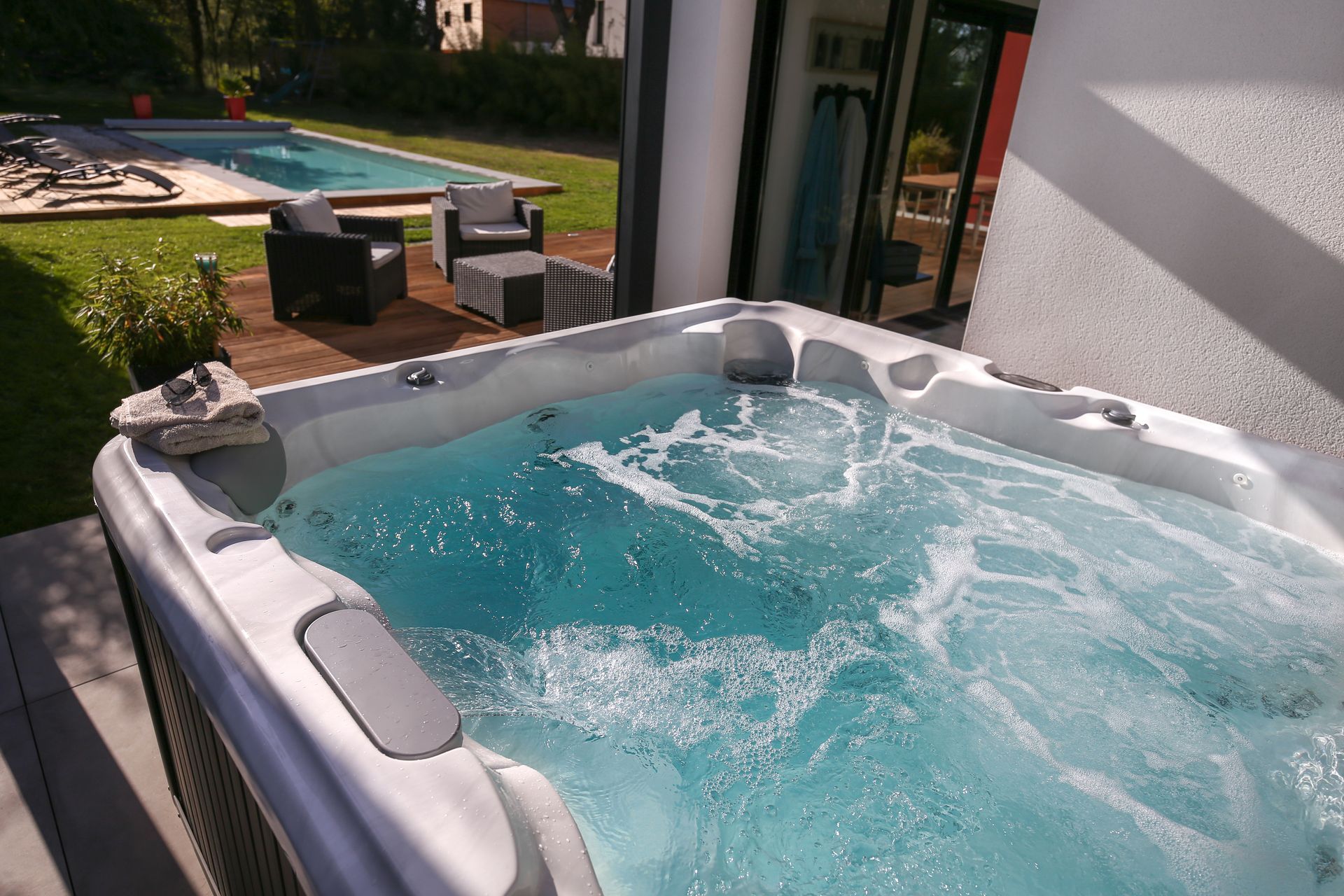 Jacuzzi dans un jardin avec eau bleue bouillonnante, à côté d'une terrasse et d'une pelouse.