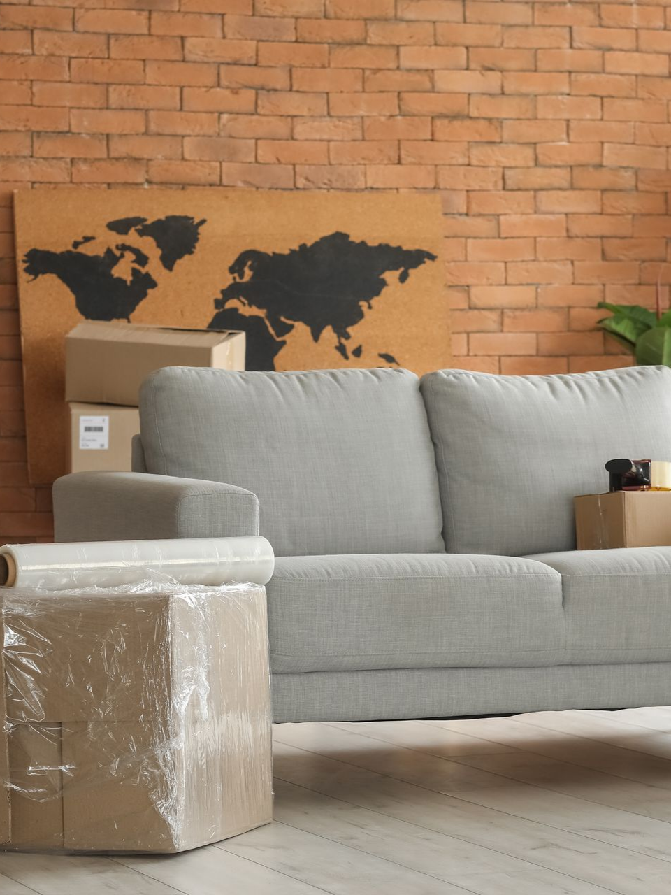 Canapé gris dans un salon aux murs de briques, avec des cartons de déménagement emballés et une carte du monde en décoration murale.