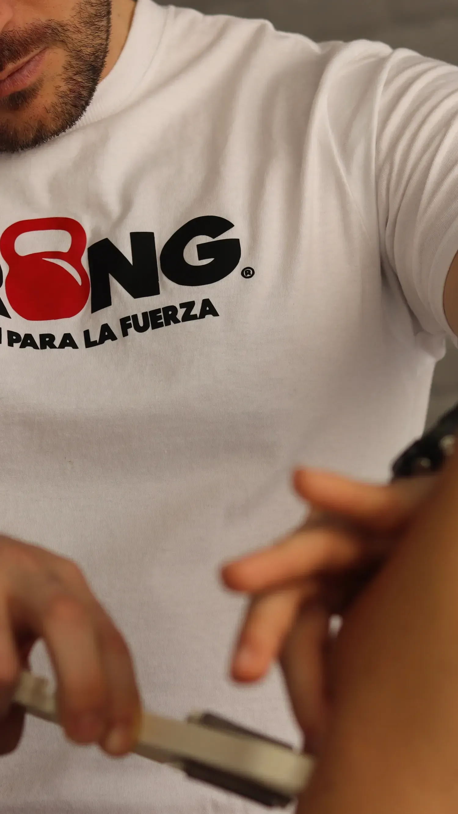 Una persona lleva una camisa blanca con el logo de una pesa rusa. Sostiene una herramienta metálica frente a otra persona.