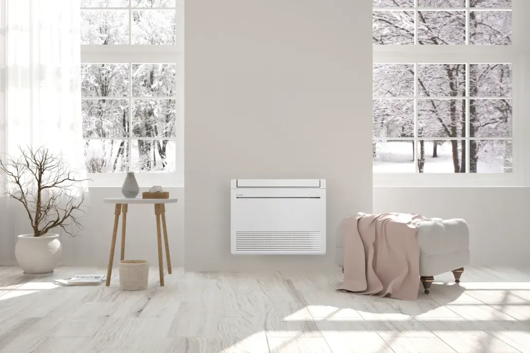 Unité de climatisation blanche fixée au mur dans une pièce aux fenêtres glaciales, avec une petite table et un pouf.