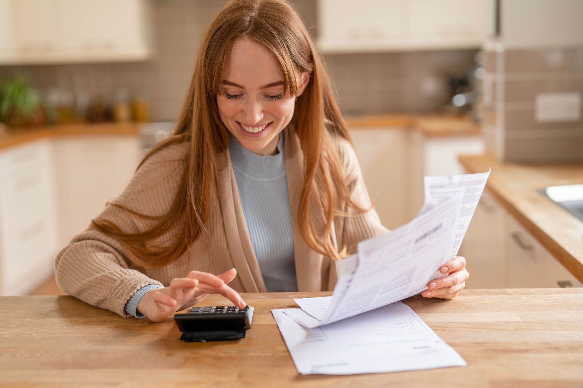 Une femme souriante, utilisant une calculatrice, révise des documents à une table de cuisine.