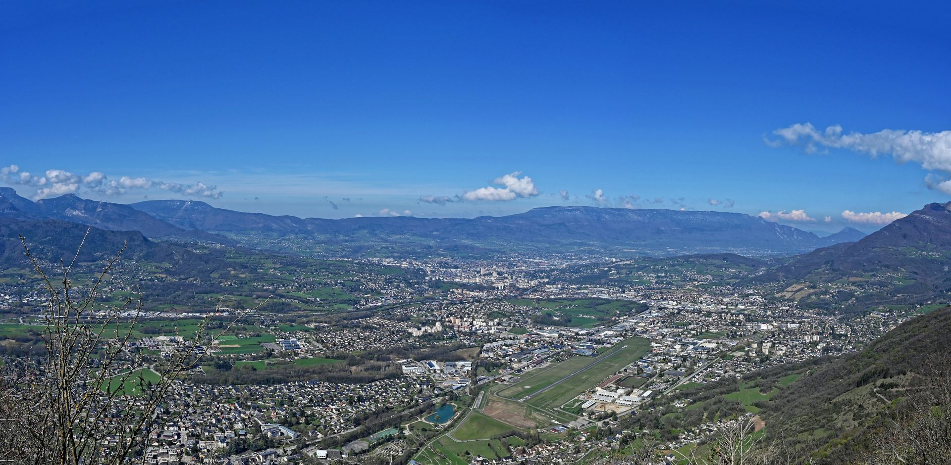 Vue panoramique d'une ville nichée dans une vallée, sous un ciel d'un bleu éclatant. Des collines et des montagnes verdoyantes entourent la ville.