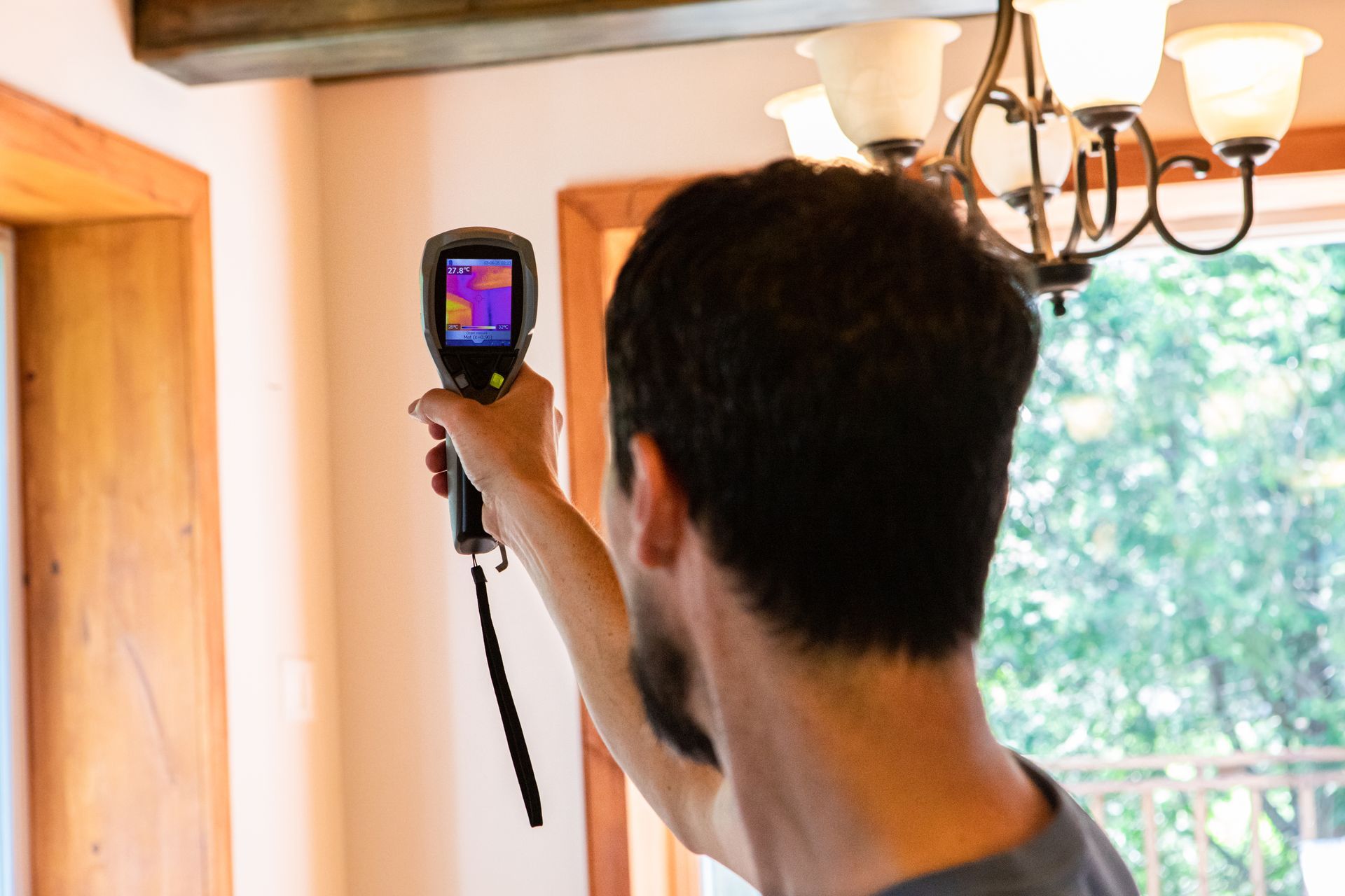 Une personne utilise une caméra thermique à l'intérieur, examinant un plafond près d'une fenêtre. L'écran affiche les variations de température.