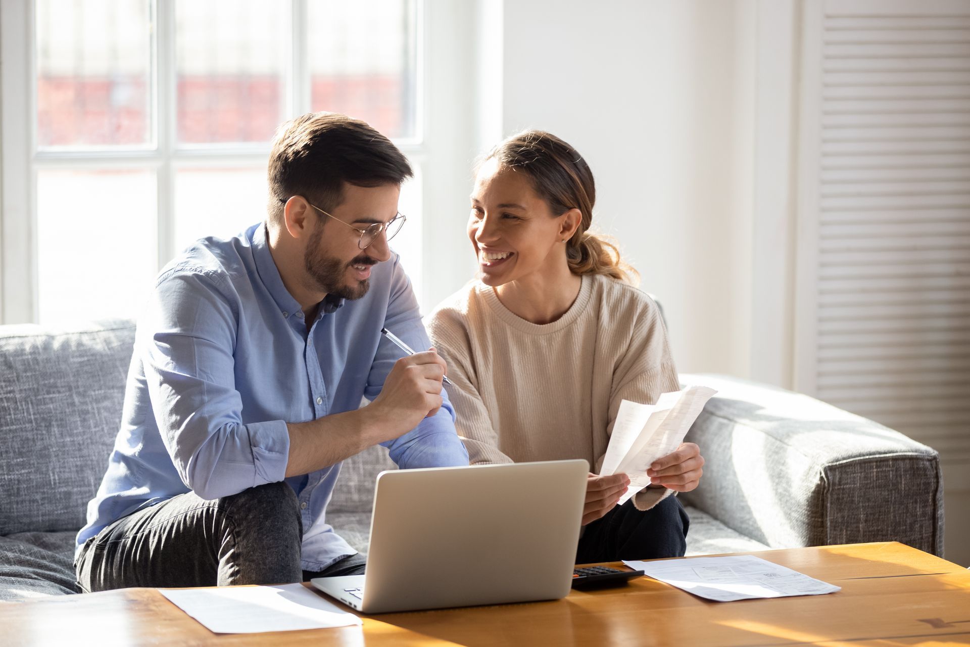 Un couple sourit en consultant des documents et en utilisant un ordinateur portable sur une table basse.