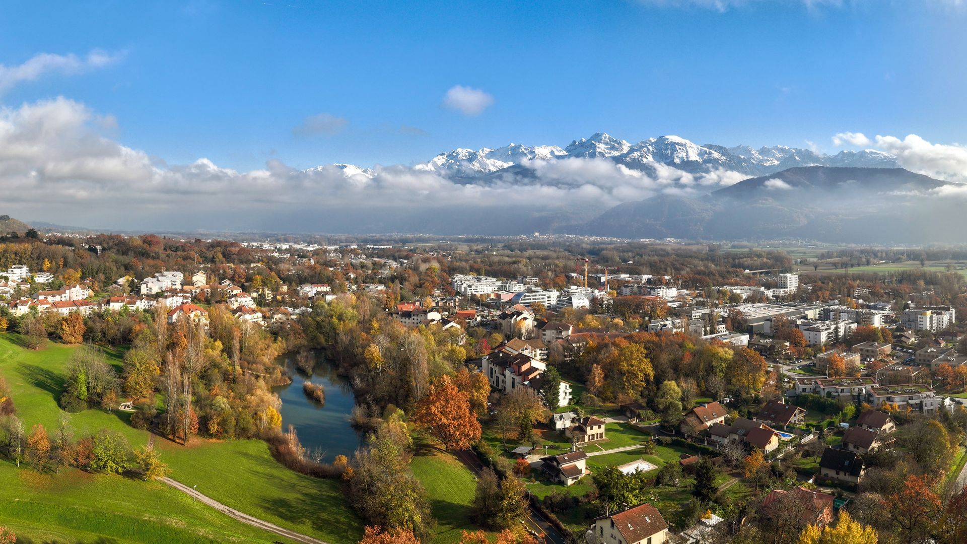 Paysage urbain avec arbres d'automne, lac et montagnes enneigées en arrière-plan, sous un ciel bleu.
