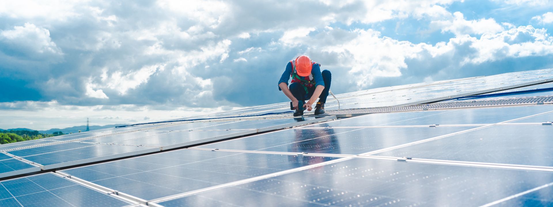 Un ouvrier portant un casque orange travaille sur un toit équipé de panneaux solaires, sous un ciel nuageux.
