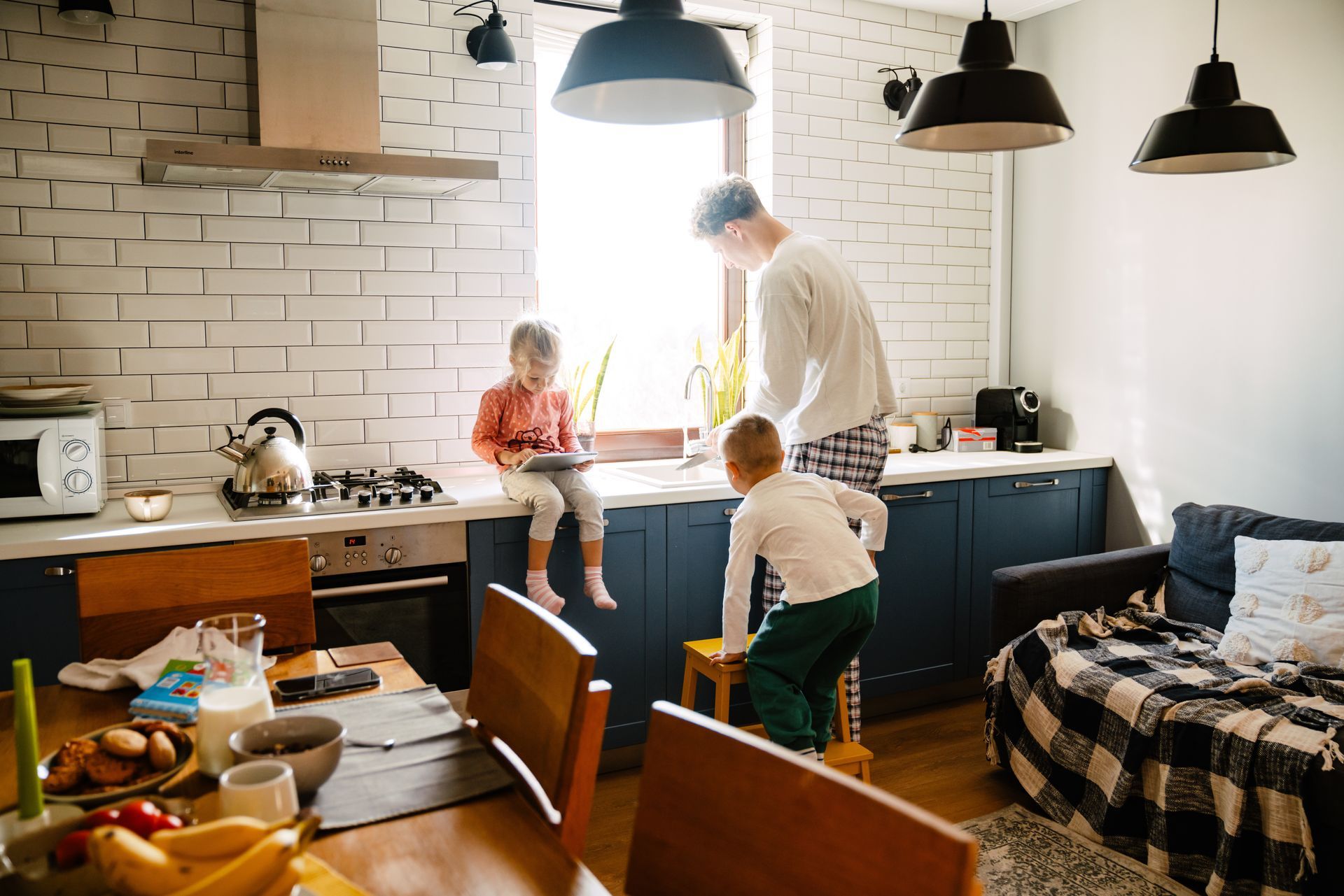 La famille dans la cuisine : un parent près de la fenêtre, deux enfants près du plan de travail et de la table où est servi le petit-déjeuner.