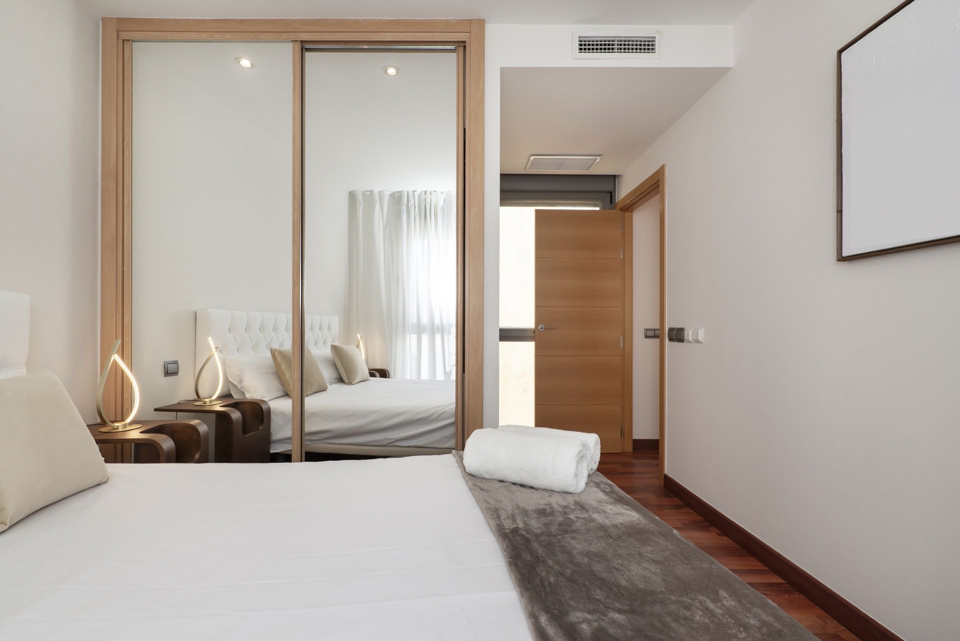 Chambre avec lit, placard à miroir, porte ouverte sur le couloir et parquet.