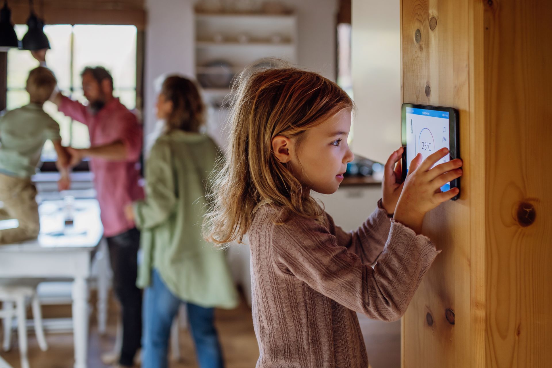 Une jeune fille utilise une tablette connectée sur un mur en bois tandis que sa famille interagit en arrière-plan.