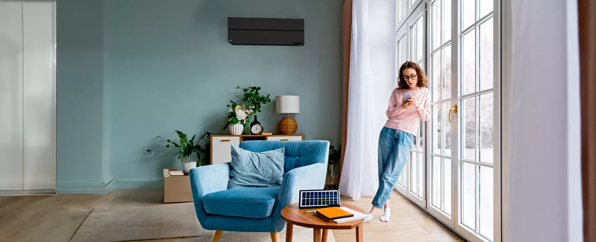 Une femme est appuyée contre une fenêtre, un téléphone à la main, dans un salon aux murs et aux meubles turquoise.