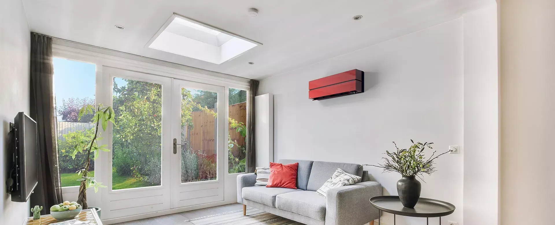 Salon lumineux avec canapé gris, coussins rouges et climatiseur rouge sur un mur blanc. Grandes portes vitrées donnant sur un jardin.