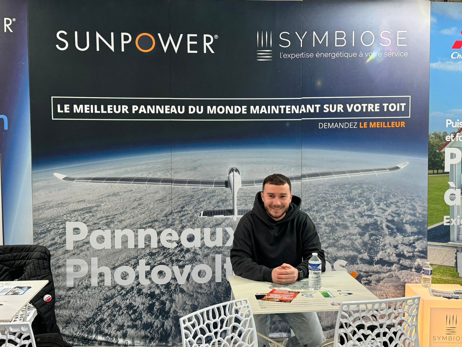 Un homme est assis à une table devant une banderole publicitaire SunPower faisant la promotion de panneaux photovoltaïques.