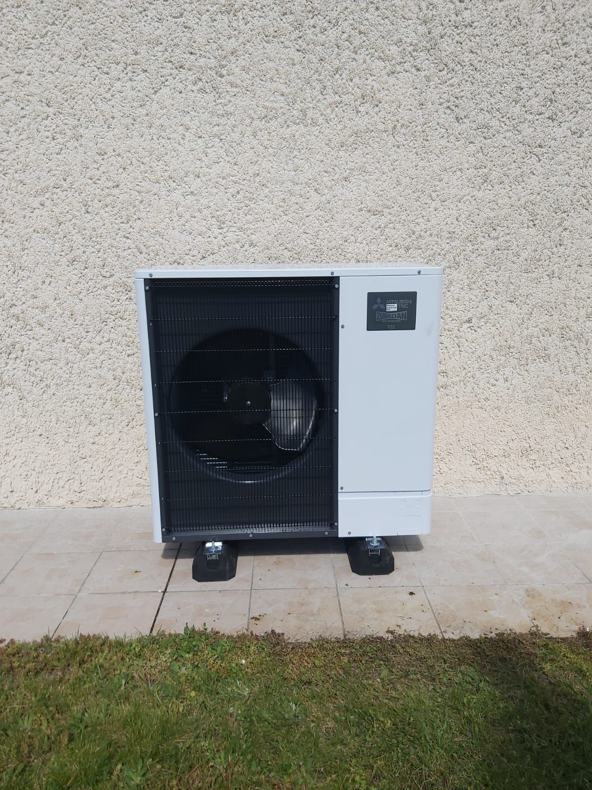 Unité de pompe à chaleur blanche sur blocs noirs, contre un mur texturé et de l'herbe.