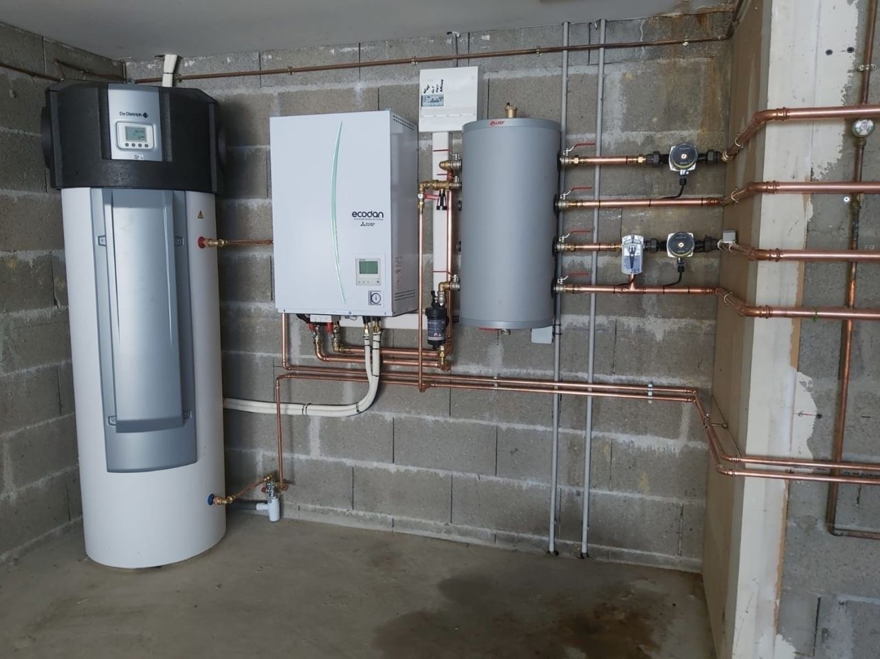 Installation du système de chauffage : ballon, pompe à chaleur, chaudière, tuyaux en cuivre contre un mur en parpaings gris.