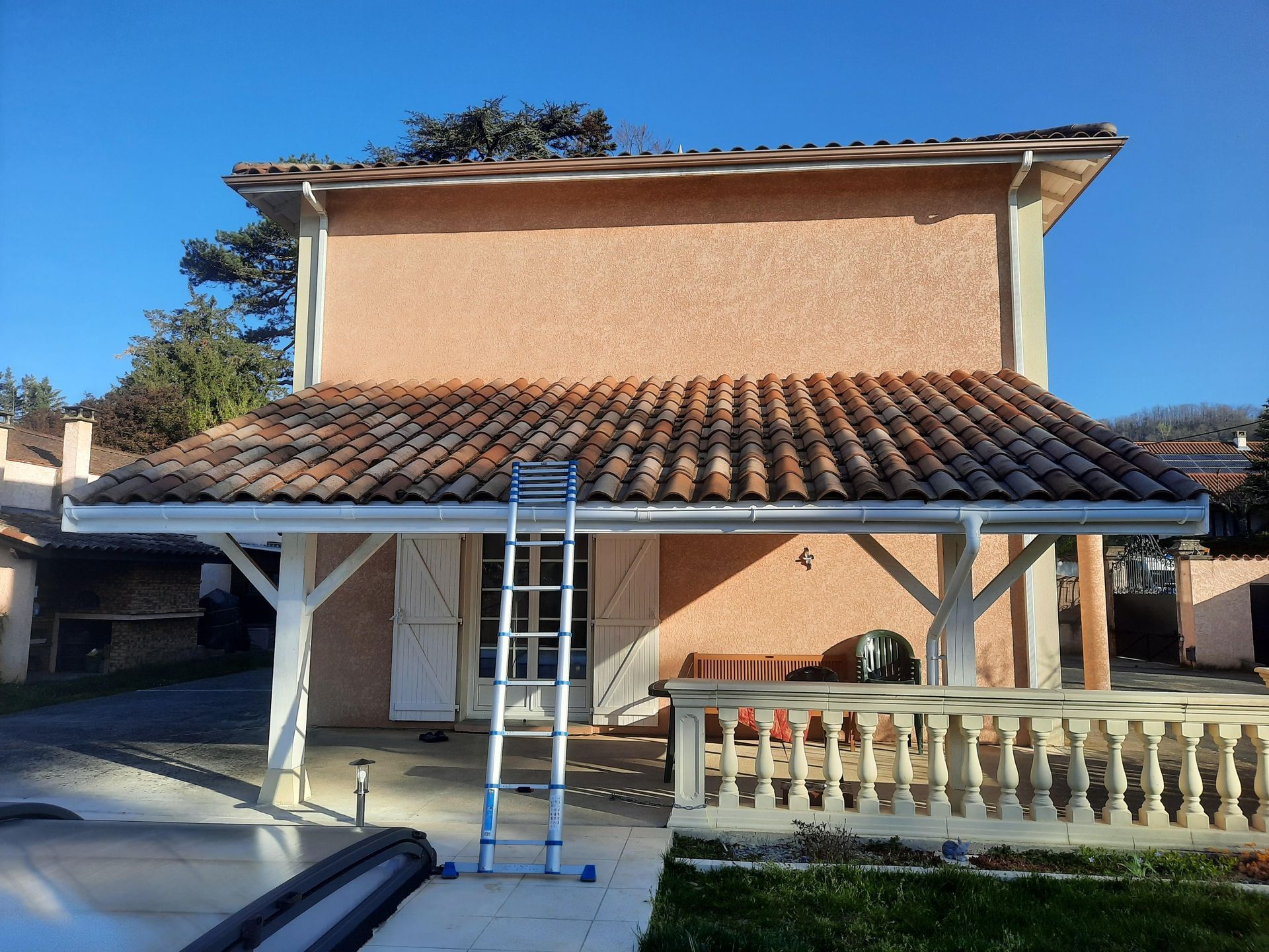 Une échelle est appuyée contre une maison au toit de tuiles et au porche couvert muni d'un système de gouttières.