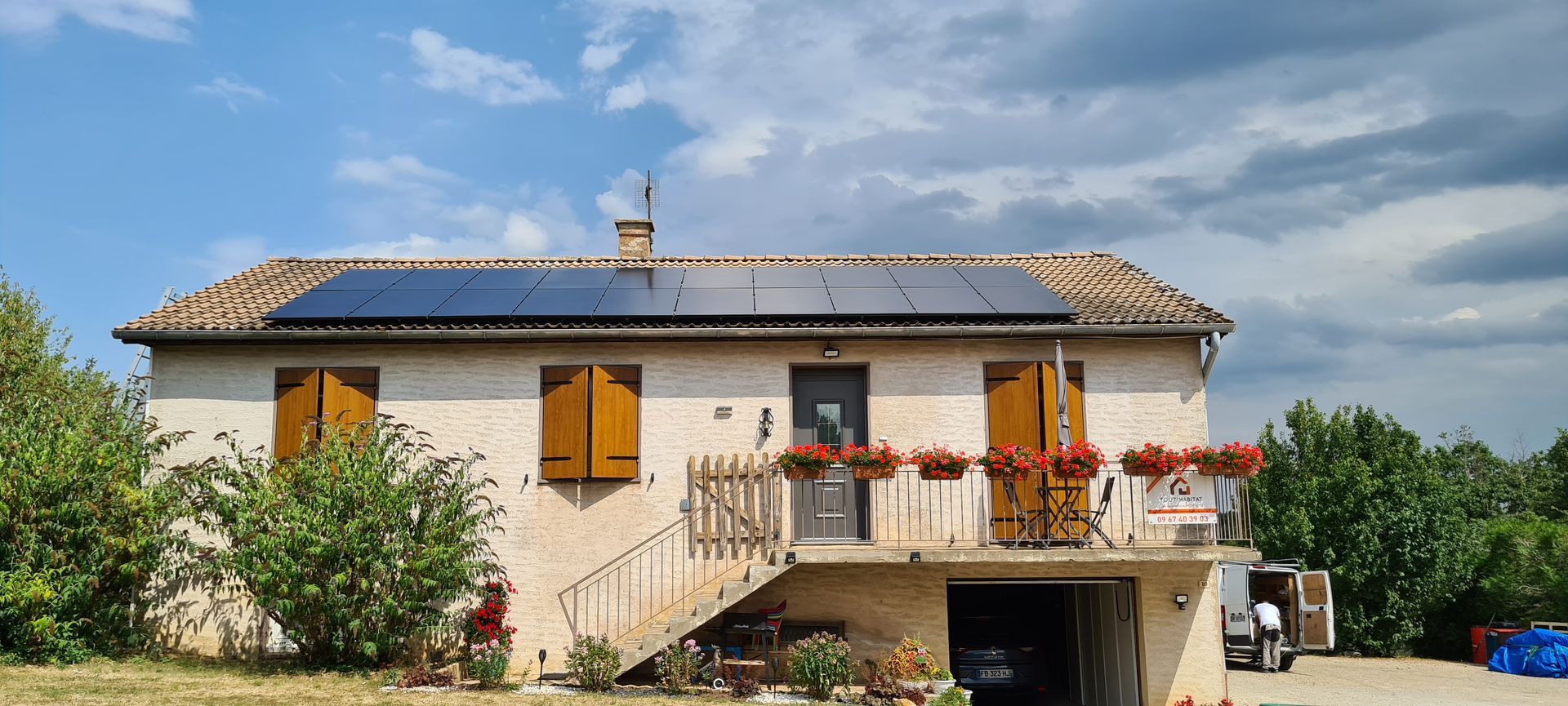 Maison avec panneaux solaires sur le toit, volets, petit balcon fleuri et ciel bleu nuageux.