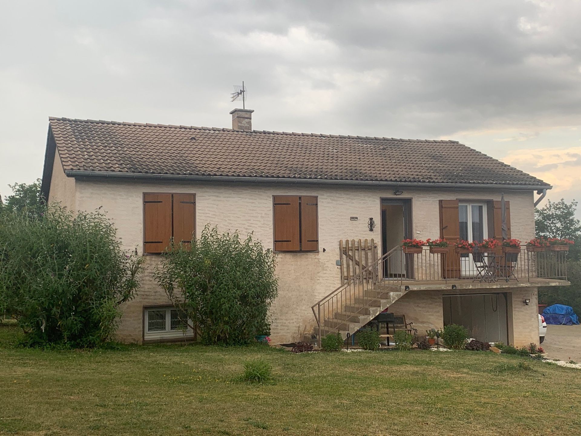 Maison beige à deux étages avec des volets marron et un balcon. Le ciel est nuageux.