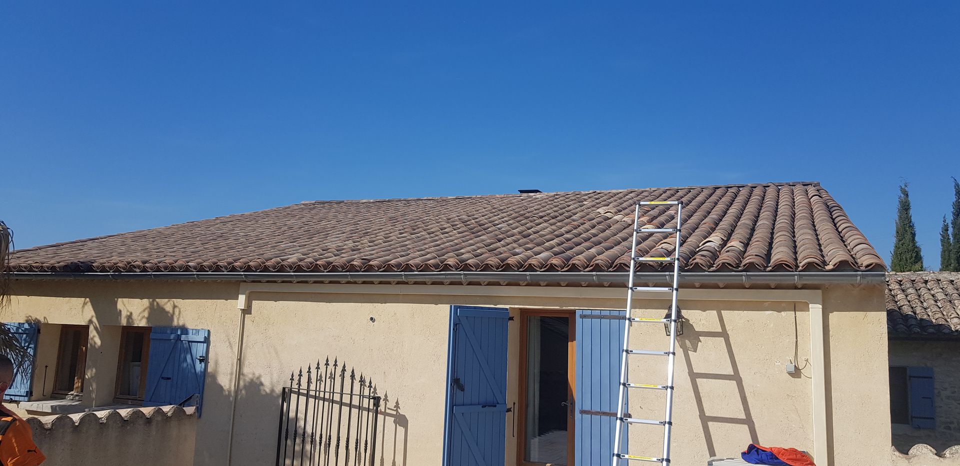 Une maison au toit de tuiles, aux volets bleus, et une échelle appuyée contre le toit par une journée ensoleillée.
