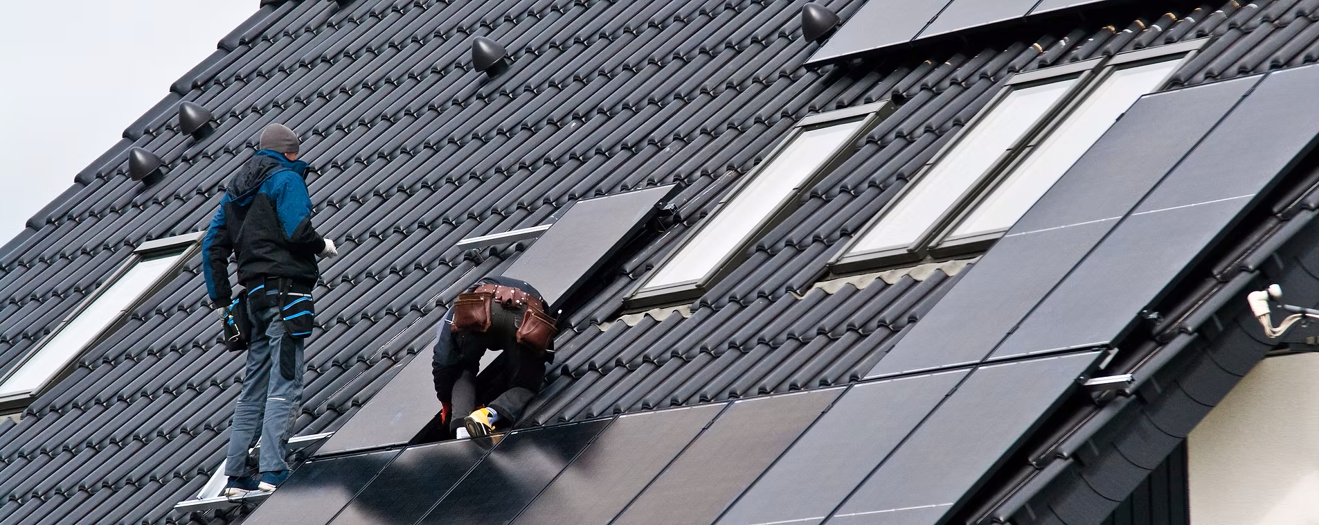 Deux ouvriers installent des panneaux solaires sur un toit de tuiles sombres, près de puits de lumière.
