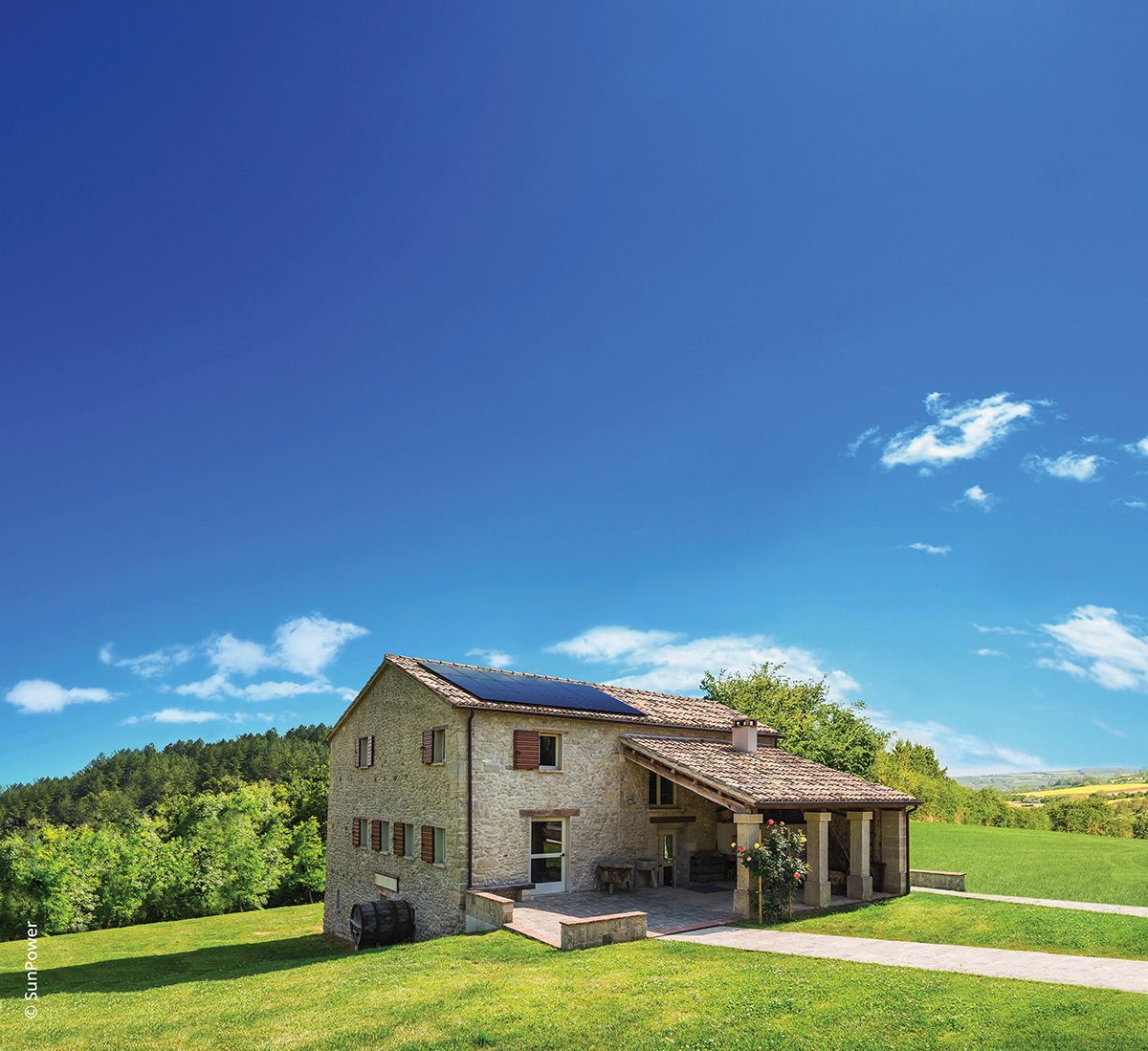 Maison en pierre avec panneaux solaires sur le toit, colline verdoyante sous un ciel d'un bleu éclatant.