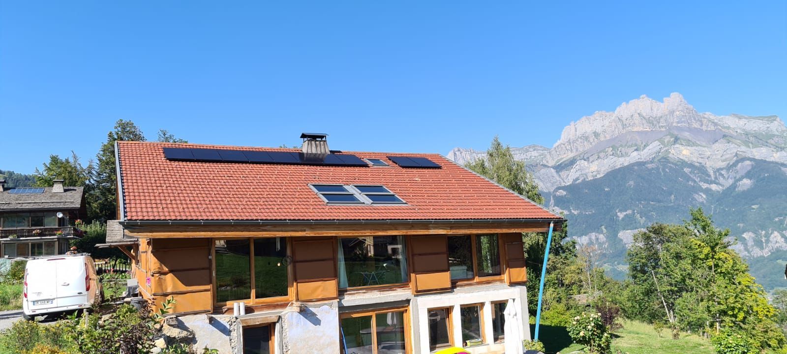 Maison en bois avec panneaux solaires sur le toit ; montagnes et arbres en arrière-plan sous un ciel bleu.