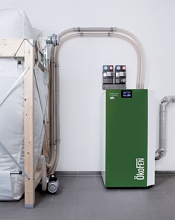 Chaudière à granulés verte et blanche avec trémie et système d'alimentation pneumatique intégrés, adossée à un mur blanc.