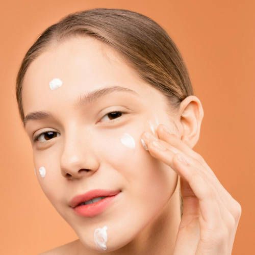 Mujer aplicándose crema hidratante en la cara, sonriendo sobre un fondo naranja.