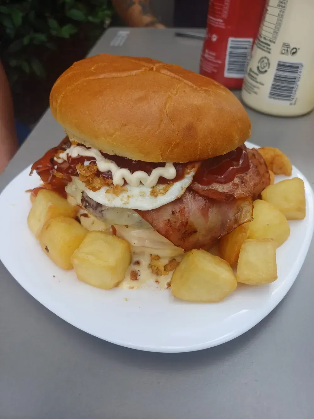 Hamburguesa con tocino, huevo frito y patatas en un plato blanco.