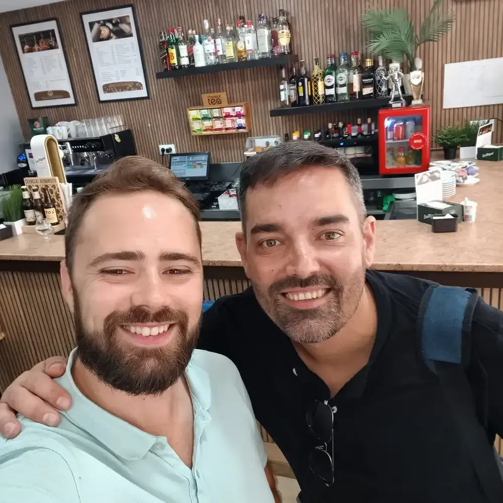 Dos hombres sonrientes, abrazados, en un café con botellas y bebidas en los estantes detrás de ellos.