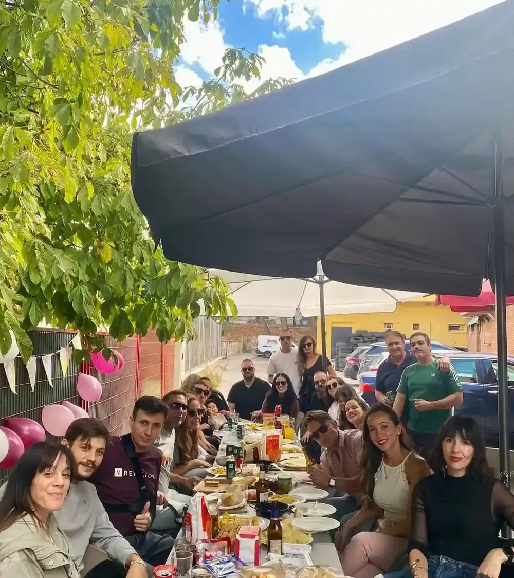 Grupo de personas en una larga mesa al aire libre bajo una sombrilla, comiendo y sonriendo.