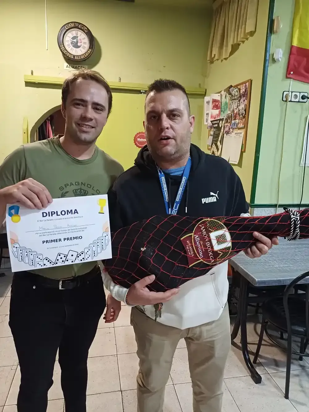 Dos hombres sostienen un diploma y un jamón curado. Uno sonríe, el otro mira hacia adelante en una habitación bien iluminada.