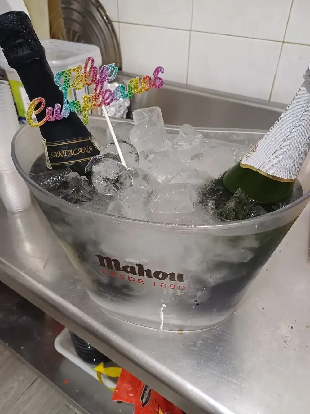 Cubo de hielo con dos botellas de champán, hielo y un adorno para tarta de cumpleaños sobre una superficie de acero.