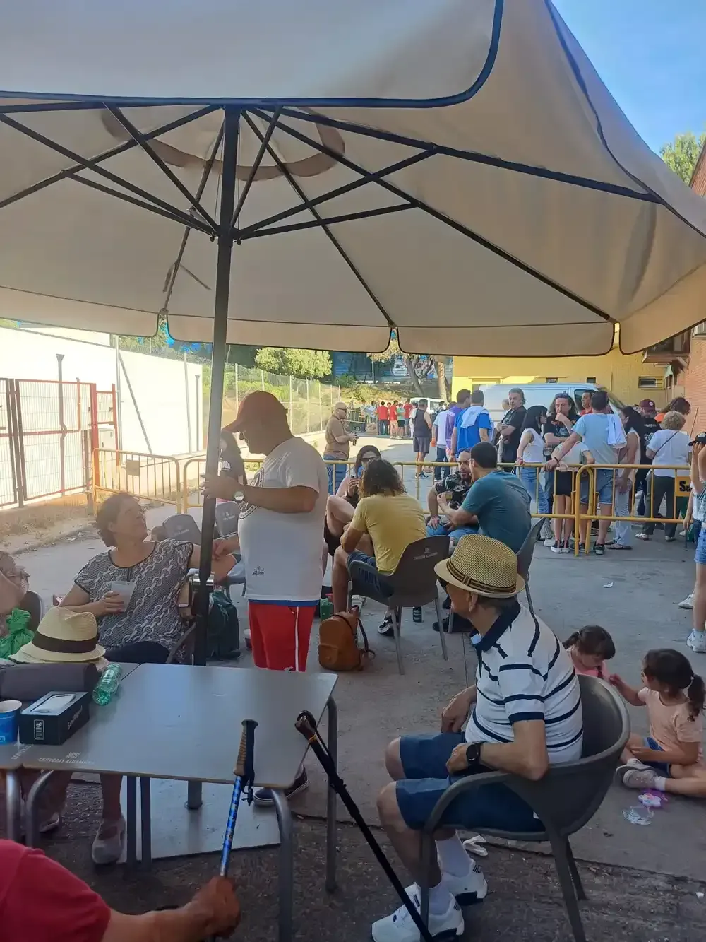 Personas bajo una gran sombrilla en una reunión al aire libre. Algunos sentados, otros haciendo fila. Día soleado.