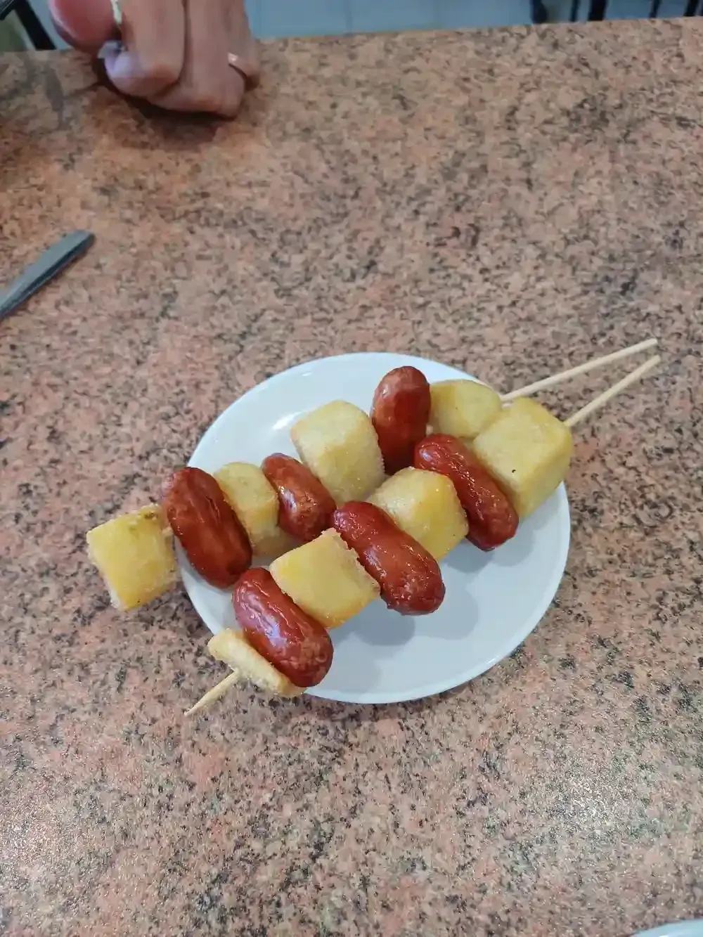 Brochetas con salchichas y patatas cortadas en cubos en un plato blanco, sobre una encimera moteada.