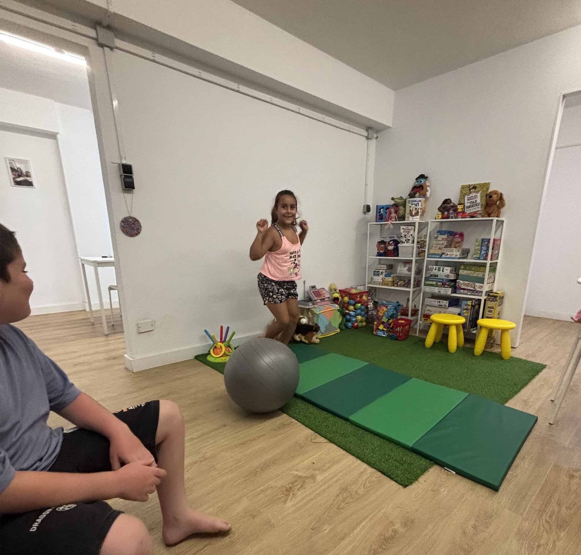 Una niña salta en un cuarto de juegos, mientras un niño está sentado cerca. Se ven juguetes, tapetes y estantes.