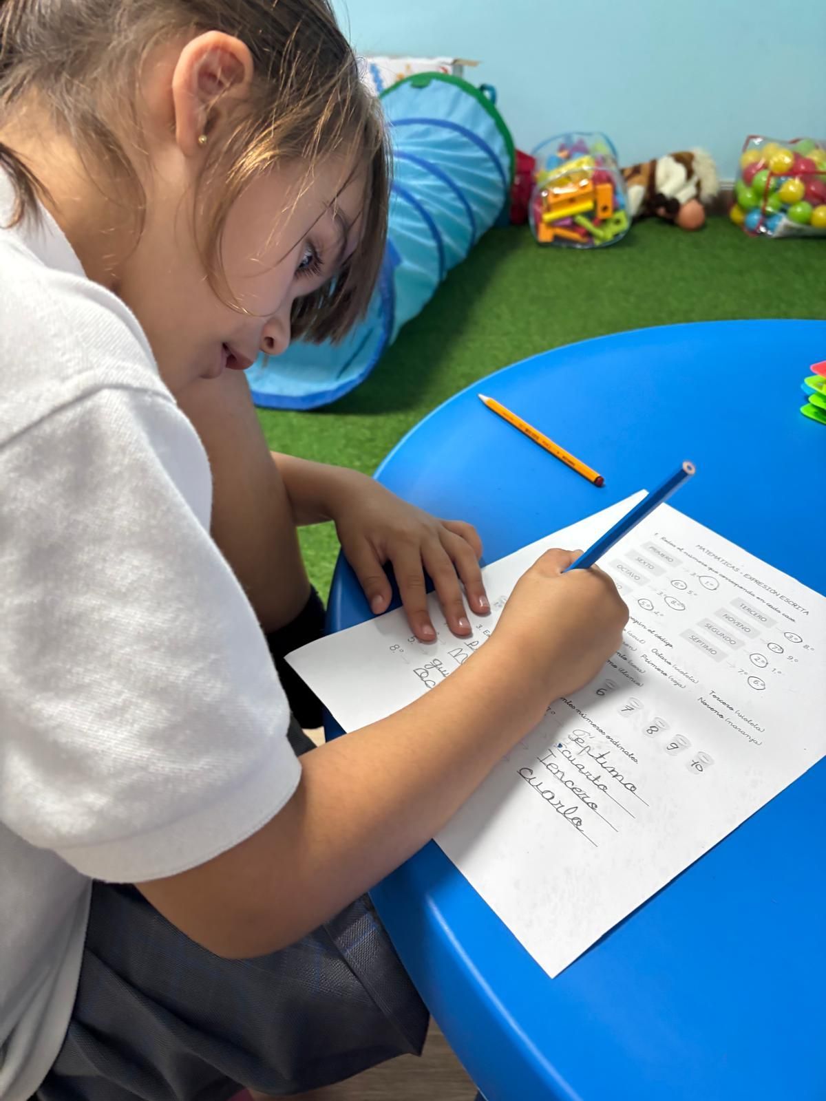 Una niña escribe en un papel en una mesa azul; al fondo hay una sala de juegos.