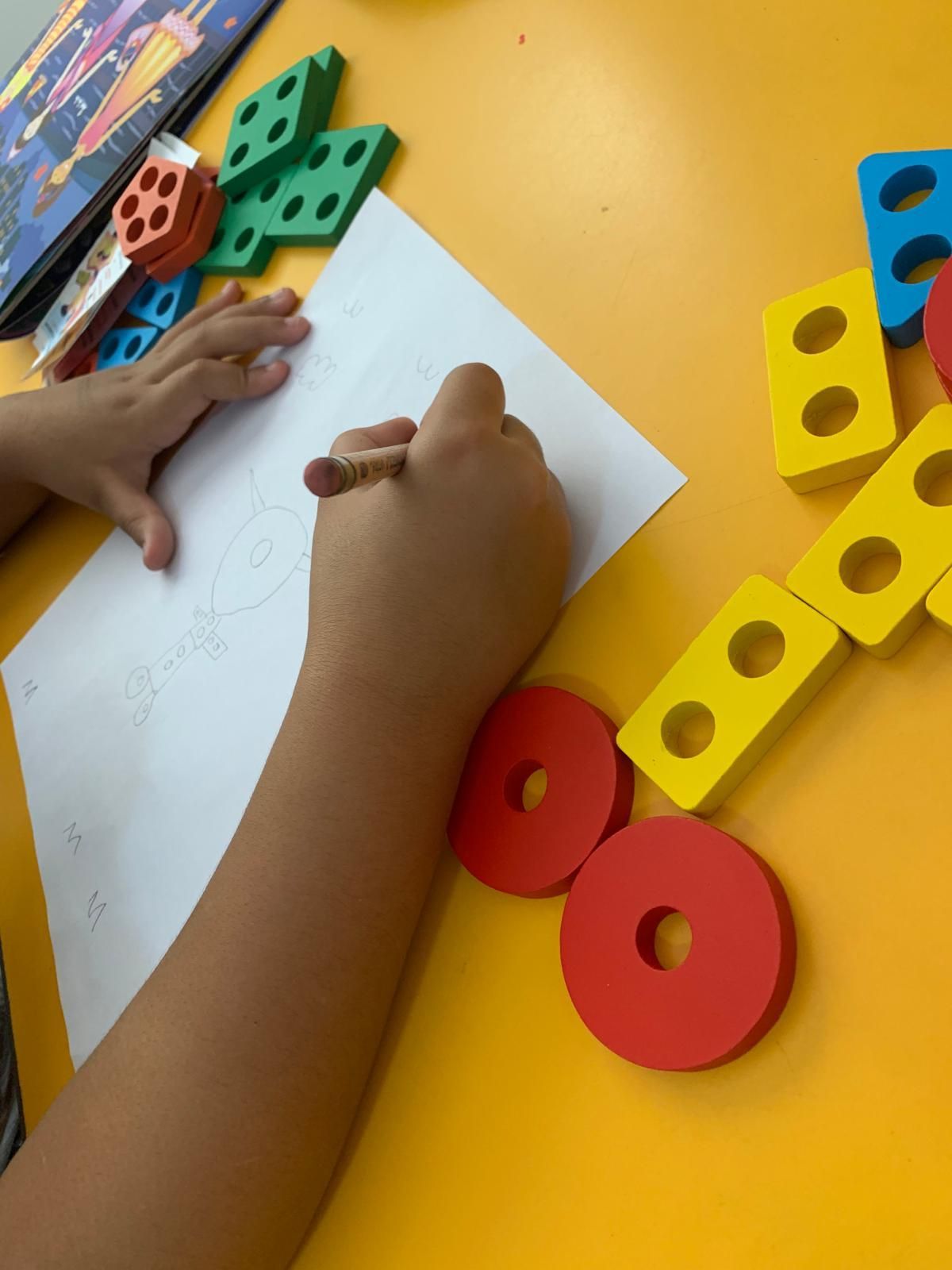 Un niño dibujando con un crayón sobre papel, rodeado de formas de madera de colores sobre una superficie amarilla.