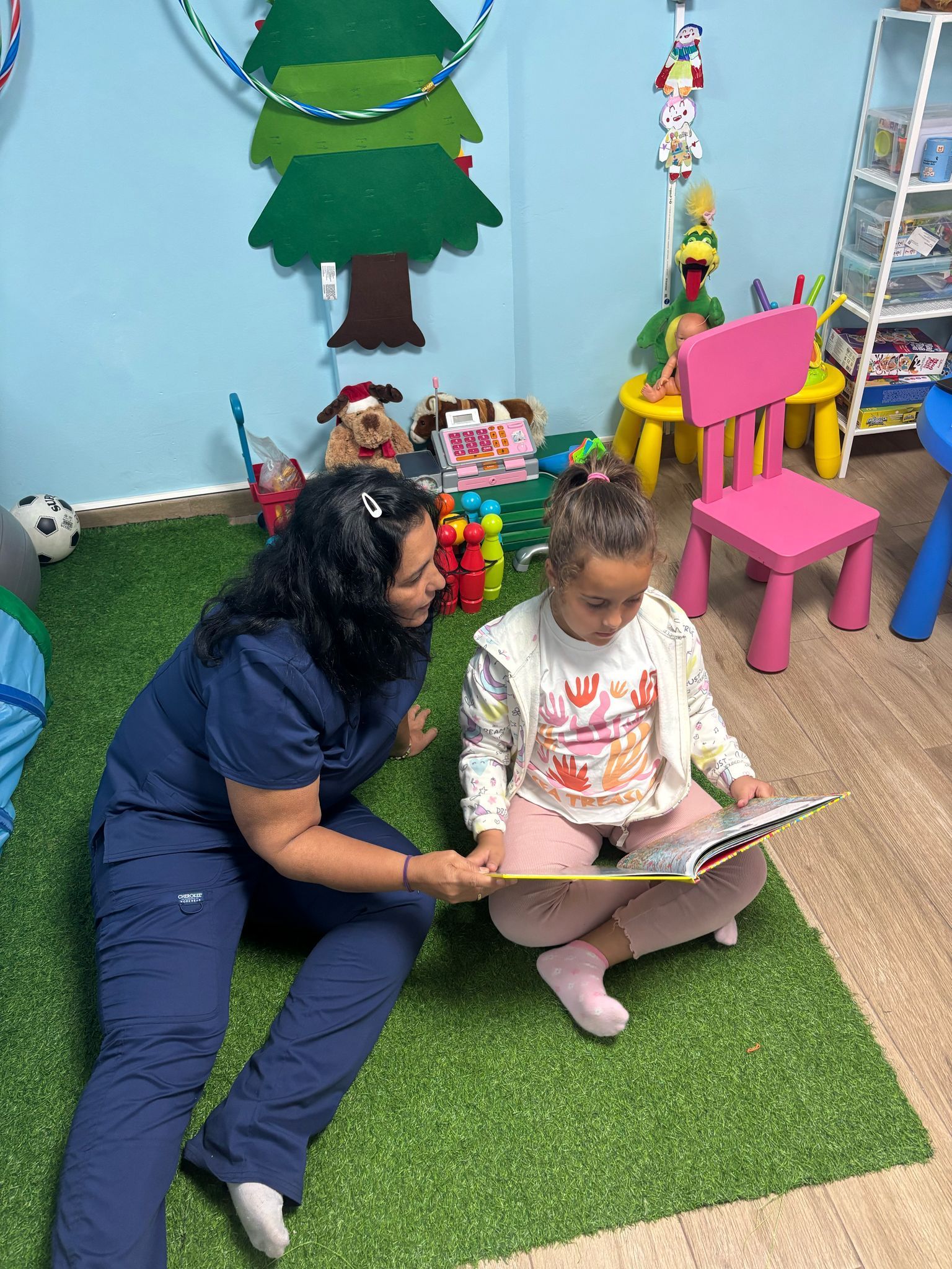 Una mujer con uniforme médico lee con un niño sentado en una alfombra verde. El niño lleva suéter y pantalones rosas.