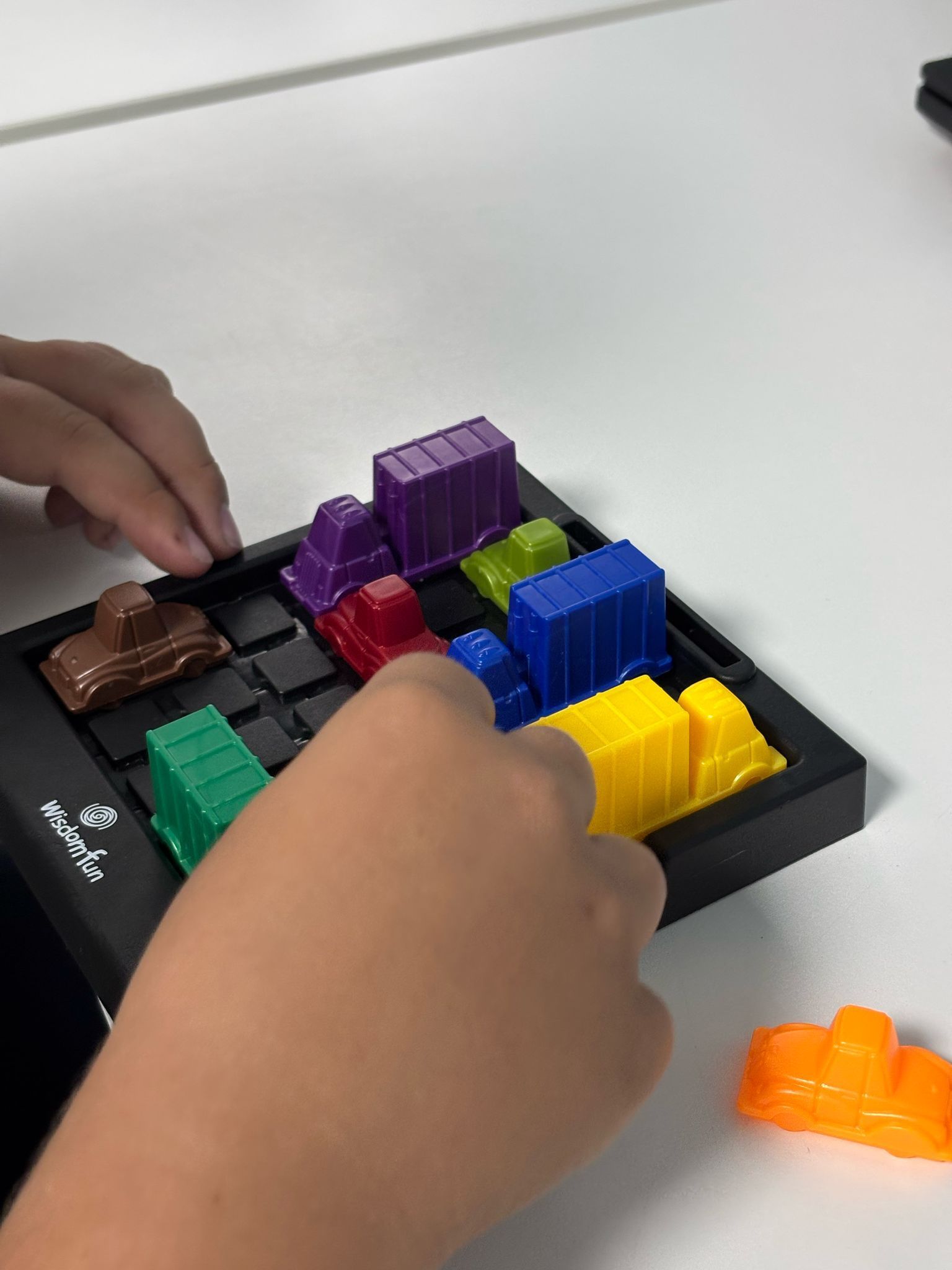 Manos jugando un rompecabezas de bloques deslizantes con vehículos coloridos que parecen juguetes sobre una cuadrícula negra.